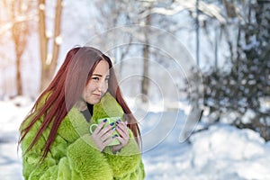 Beautiful cute pretty redhead female teenager in a green fur coat drinking coffee in the sunny winter city park.