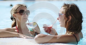 Beautiful cute women swims in a pool with clear blue water and drinks a colorful blue red yellow cocktail.