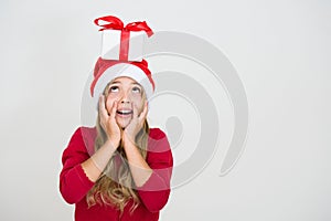Beautiful cute young girl in red dress and santa hat with gift on head looking above. Excited surprised child with present