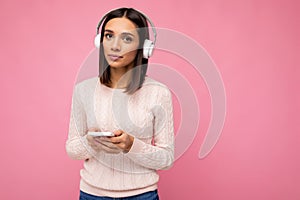 Beautiful cute young woman wearing pink sweater isolated over pink background wall wearing white bluetooth wireless