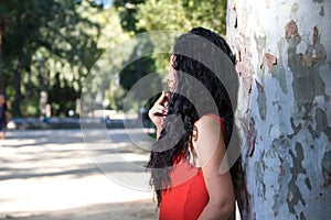 A beautiful, dark-haired, curly-haired adult woman with latin features is leaning against a giant tree. The woman covers her eye