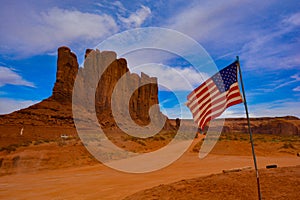 A beautiful day in Monument Valley, cloudy blue sky over camel butte and American flag in front. Red rocks and dirt road in Utah