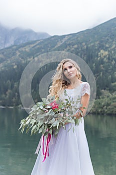 Beautiful delicate girl in the air blue bride wedding dress with luxurious curls in the mountains near the lake with a wedding bou