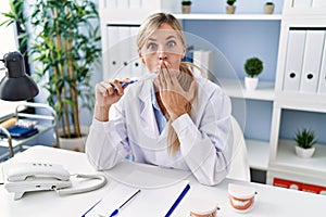 Beautiful dentist woman explaining how to brush teeth with toothbrush covering mouth with hand, shocked and afraid for mistake