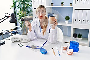 Beautiful dentist woman holding denture and toothbrush puffing cheeks with funny face