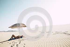 Beautiful desert dunes beach and caucasian female lay down enjoying a sunbath under a coloured umbrella - concept of summer