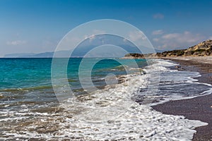 A beautiful deserted sand and pebble beach with foamy waves surf and mountains and a blue sky with white clouds