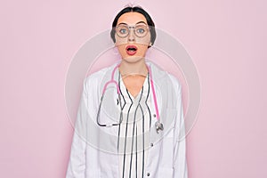 Beautiful doctor woman with blue eyes wearing coat and stethoscope over pink background afraid and shocked with surprise