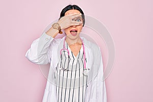 Beautiful doctor woman with blue eyes wearing coat and stethoscope over pink background peeking in shock covering face and eyes