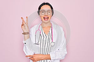 Beautiful doctor woman with blue eyes wearing coat and stethoscope over pink background smiling with happy face winking at the