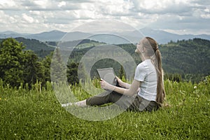 Beautiful dreaming girl with a laptop sitting on green grass on a background of mountains