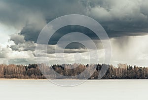 Beautiful early spring landscape with frozen lake, forest on the horizon and dramatic sky with grey clouds
