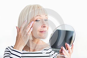 Beautiful elderly woman applying anti-wrinkle cream, holding mirror