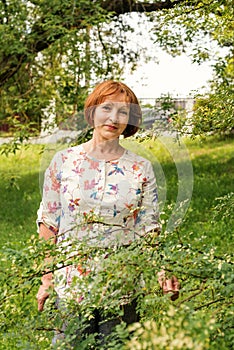 A beautiful elderly woman in flaxen clothes stands tranquilly in her green garden