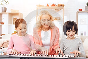 Beautiful elderly woman teaches small grandchildren to play synthesizer.