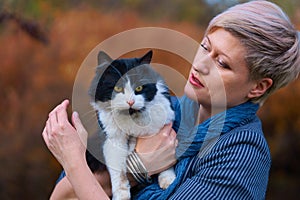 Beautiful elegant blond woman playing with cat in autumn city park, dressed in blue jeans and jacket