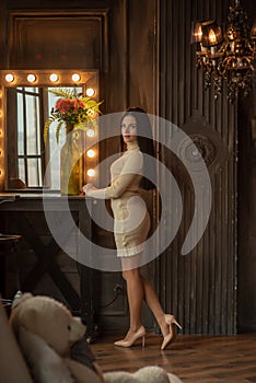 Beautiful elegant brunette is standing in front of a tall make-up mirror. A girl is standing at the cosmetic table