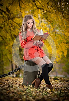 Beautiful elegant girl with orange coat reading sitting on a stump autumnal park. Young pretty woman with blonde hair reading