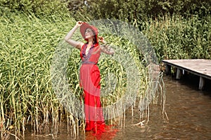 A beautiful elegant lady in a red hat and a long red dress walks along the water along the shore with reeds. emotional portrait of