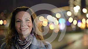 beautiful and elegant middle-aged woman is walking in city in night, looking to camera and smiling