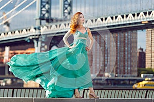 Beautiful elegant red haired woman standing near Manhattan Bridge in New York City wearing long green dress.