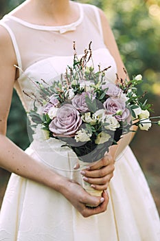 Beautiful elegant summer wedding bouquet of lilac roses and wildflowers in the hands of the bride in white wedding dress