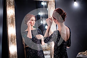 Beautiful, elegant woman in black evening dress stands next to dark high mirror