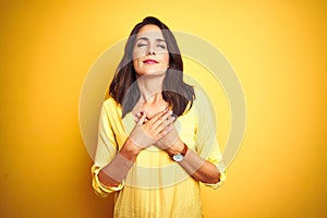 Beautiful elegant woman standing over yellow isolated background smiling with hands on chest with closed eyes and grateful gesture