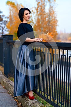 Beautiful elegant woman standing and posing with bouquet of yellow leaves at riverside parkway in autumn city park