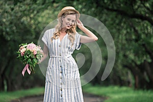 beautiful elegant woman in straw hat with bouquet of flowers standing on path