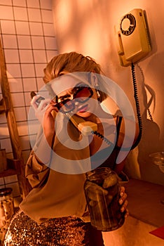 beautiful elegant woman in sunglasses talking on retro telephone and holding jar with pickled cucumbers in kitchen