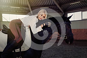 Beautiful elegant young blonde girl lies on a her black horse dressing uniform competition white blouse shirt and brown pants.