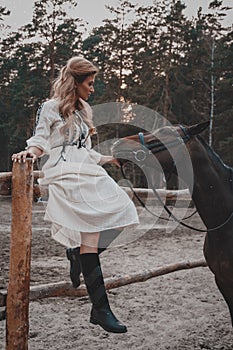 Beautiful and elegant young woman wearing the dress is stroking the horse on the ranch