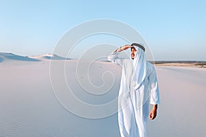 Beautiful Emirate Sheikh looks out in distance caravan of camels, standing among wide desert on hot day.