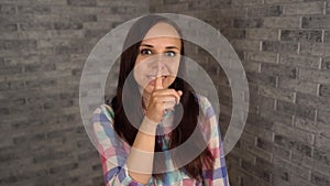 A beautiful emotional woman in a plaid shirt is doing silencing gesture with index finger at her lips on a gray brick