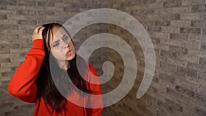 A beautiful emotional woman in a red hoodie is scratching her head in thought on a brick background.