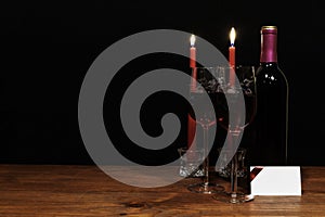 Beautiful etched wine glasses and bottle of red wine, lighted by red candles, on wooden table with name tag on dark background