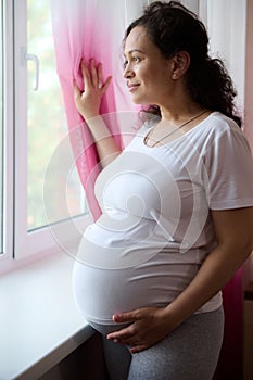 Beautiful ethnic pregnant woman, expectant mother dreamily looking out the window, touching her belly in late pregnancy