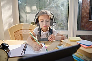 Beautiful European child schoolgirl studies with video online lesson on a digital device at home. She learns the writing