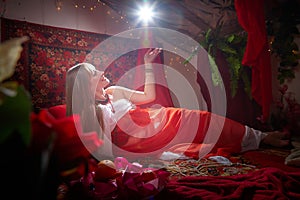 Beautiful European girl looking like Arab woman in red room with rich fabrics and carpets in sultan harem. Photo shoot