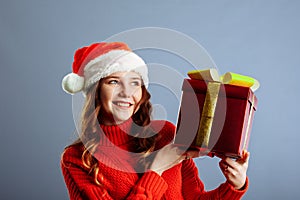 Beautiful european lady in Santa hat is holding presents, looking at camera and smiling, isolated on gray background