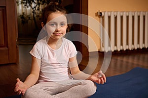 Beautiful European little girl in sports uniform, smiling looking at camera while sitting in lotus position on yoga mat.