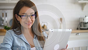 Beautiful European woman communicating using tablet sitting at table in home kitchen.