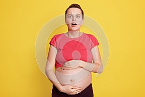 Beautiful European woman expecting baby, touching pregnant belly, looks afraid and shocked, has surprise and amazed expression,