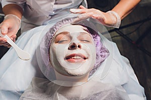 Beautiful european woman having facial skin procedure by applying a white rejuvenation mask by cosmetologist.