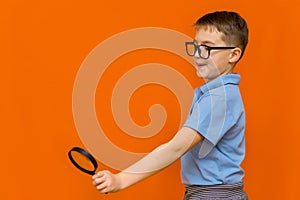 Beautiful european young boy with glasses holding and looking through magnifying glass.Orange wall