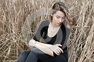 Beautiful european young woman in vintage jeans in black fashionable t-shirt sits among autumn dry grass in a field.