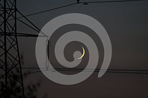 Beautiful evening view of crescent Moon near insulator of electricity transmission pylon tower and power lines seen in Ticknock