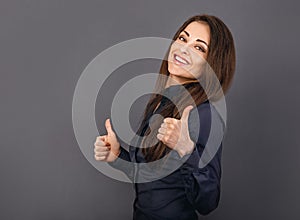 Beautiful excited energetic smiling business woman showing fingers thumb up, the ok sign in blue shirt on grey background. Closeup