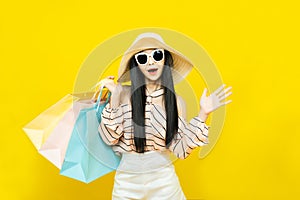 Beautiful excited happy young shopaholic asian woman wearing sungalsses and floppy hat posing isolated over yellow background